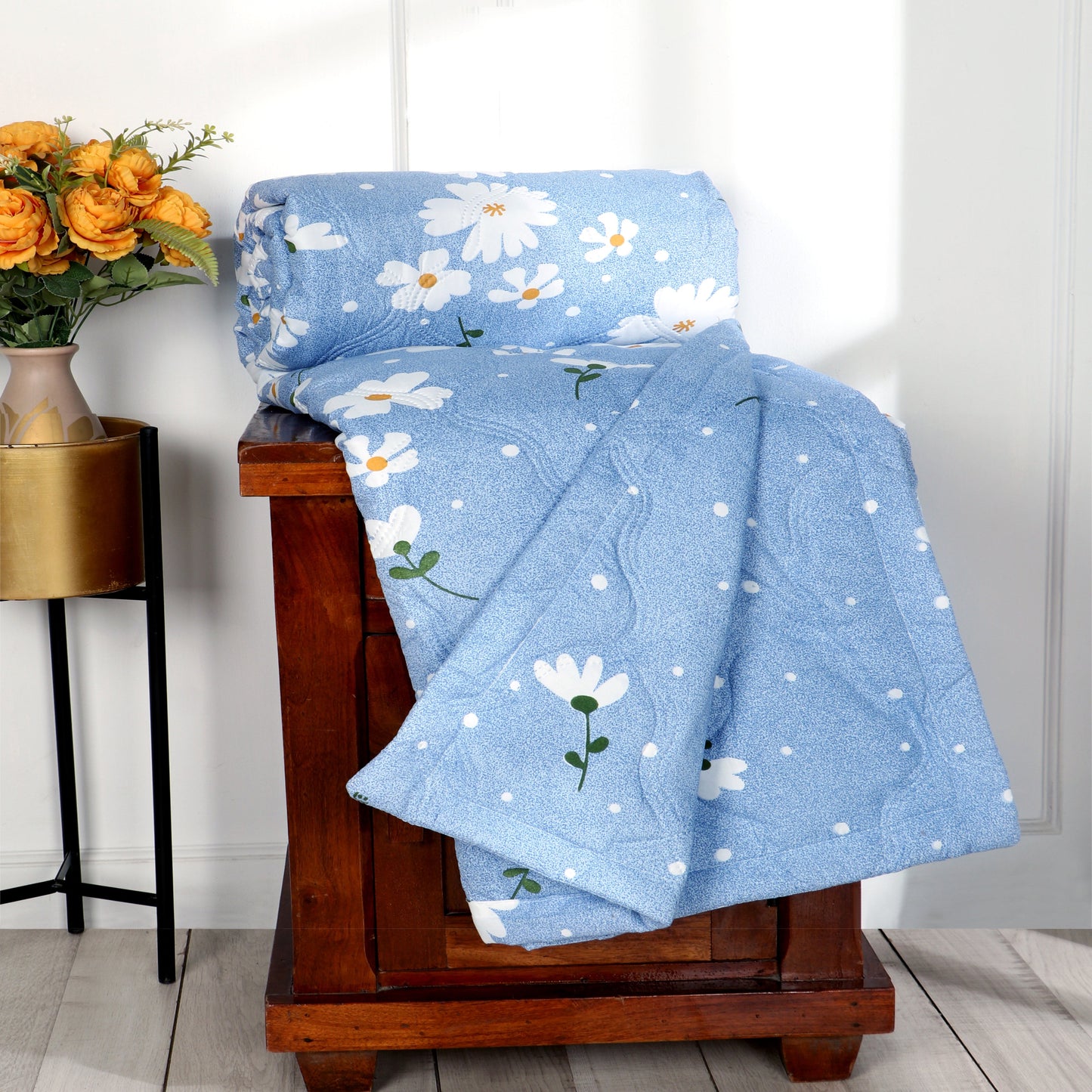 Blue floral quilt draped over a wooden bench with a side table and flowers in the background.